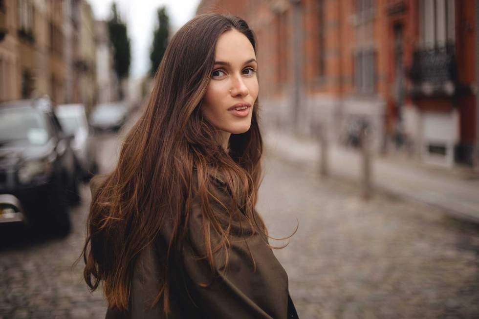 Calm smiling long hair brunette woman cross road, while walking outdoor in old town. Thoughtful woman wear green trench coat. Fashion style outfit.