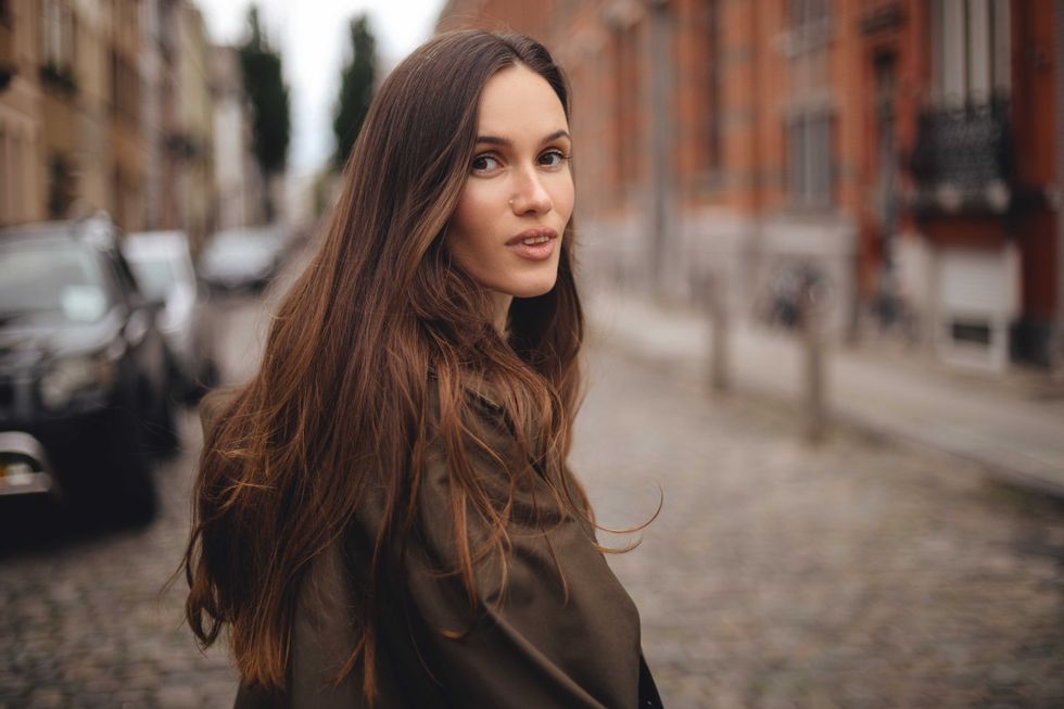 Calm smiling long hair brunette woman cross road, while walking outdoor in old town. Thoughtful woman wear green trench coat. Fashion style outfit.