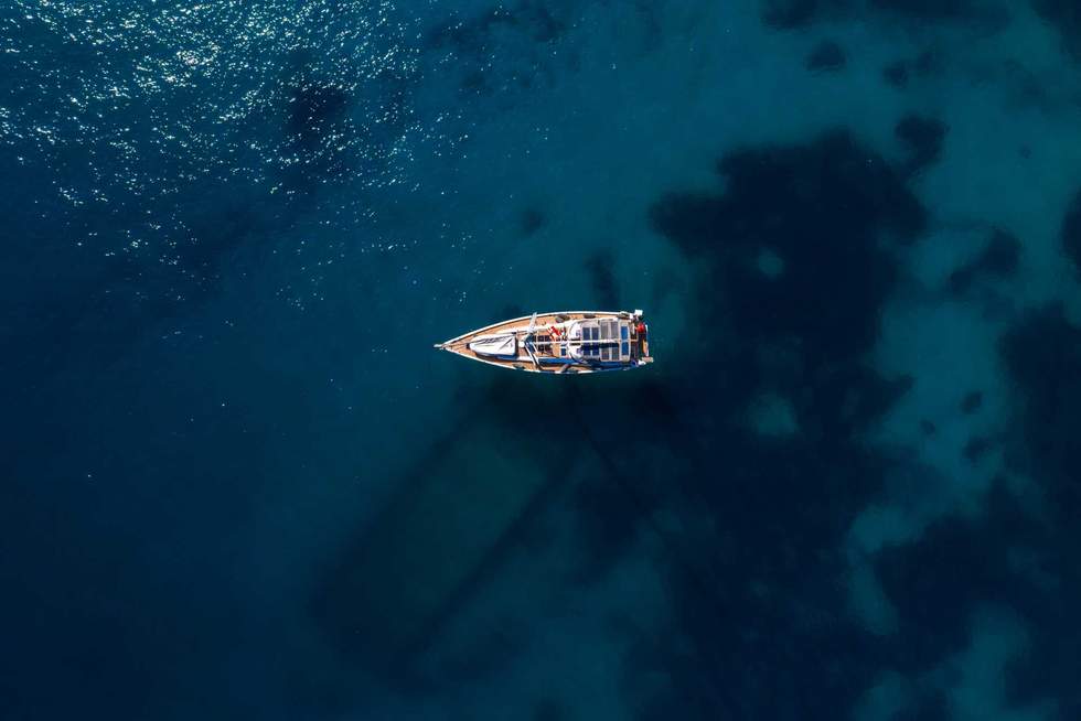 Directly above, a single sailboat rests on deep blue water. Sun glints on the sea, reefs tint patches, and the hull casts a long diagonal shadow.