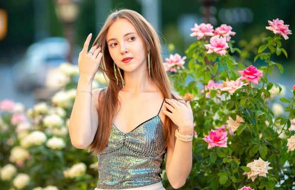 Elegant young woman posing among blooming pink roses in a summer garden. Romantic beauty portrait with long hair, stylish outfit and natural floral background, lifestyle concept.