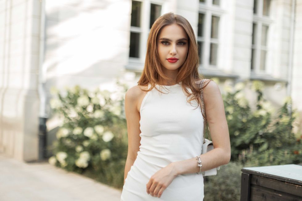 Fashionable beautiful woman in a white elegant dress with a handbag walks on the street