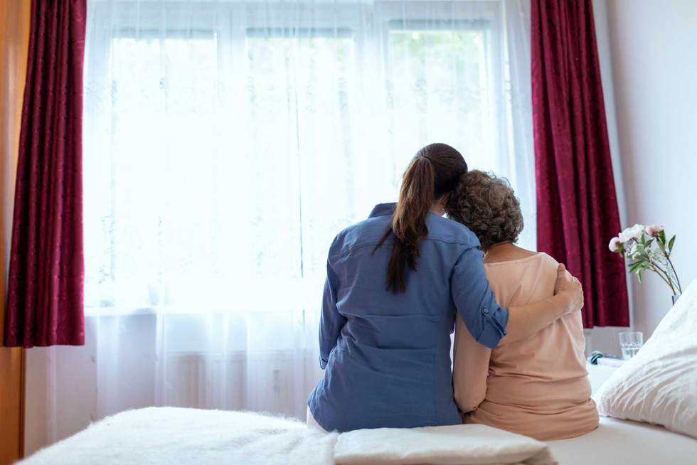 Female Home Nurse Hugging Elderly Woman on Bed. Back Wiev of Female Nurse With Her Arm Around Elderly Patient Shoulder.