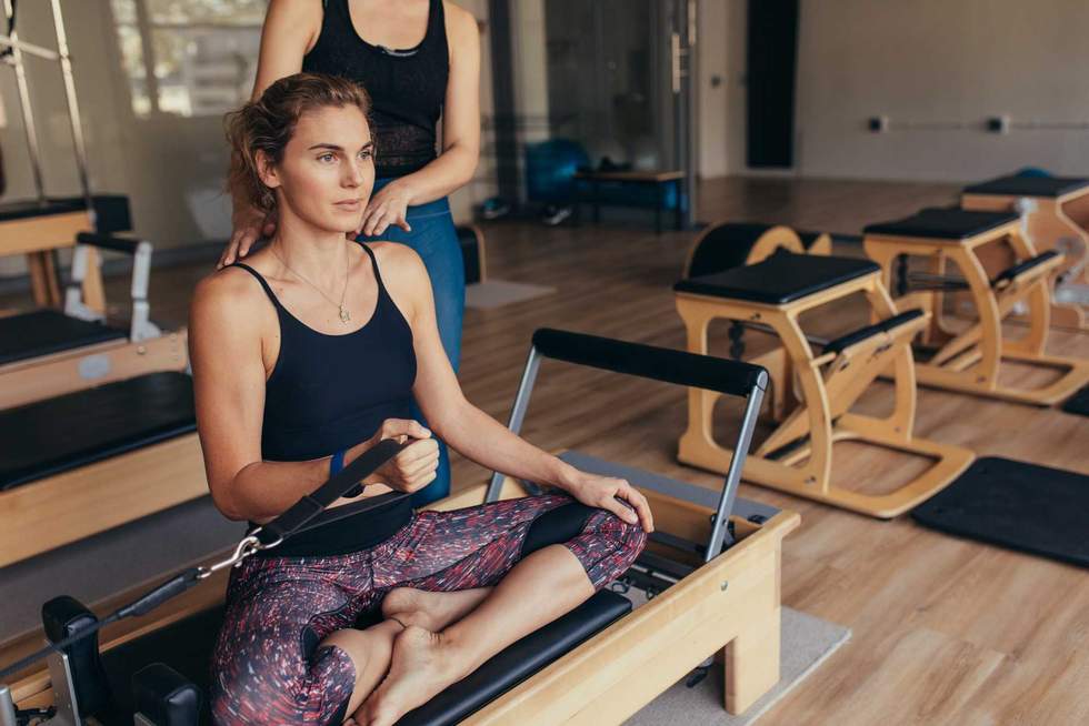Fitness women sitting with legs crossed on a pilates training machine and pulling the stretch band. Pilates trainer guiding a woman at the gym doing strength training.