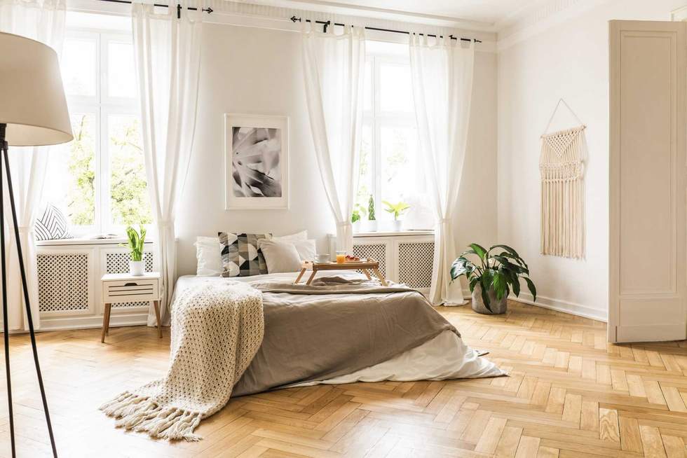 Framed poster on a white wall above a cozy double bed with beige sheets and breakfast tray in a spacious, sunny bedroom interior