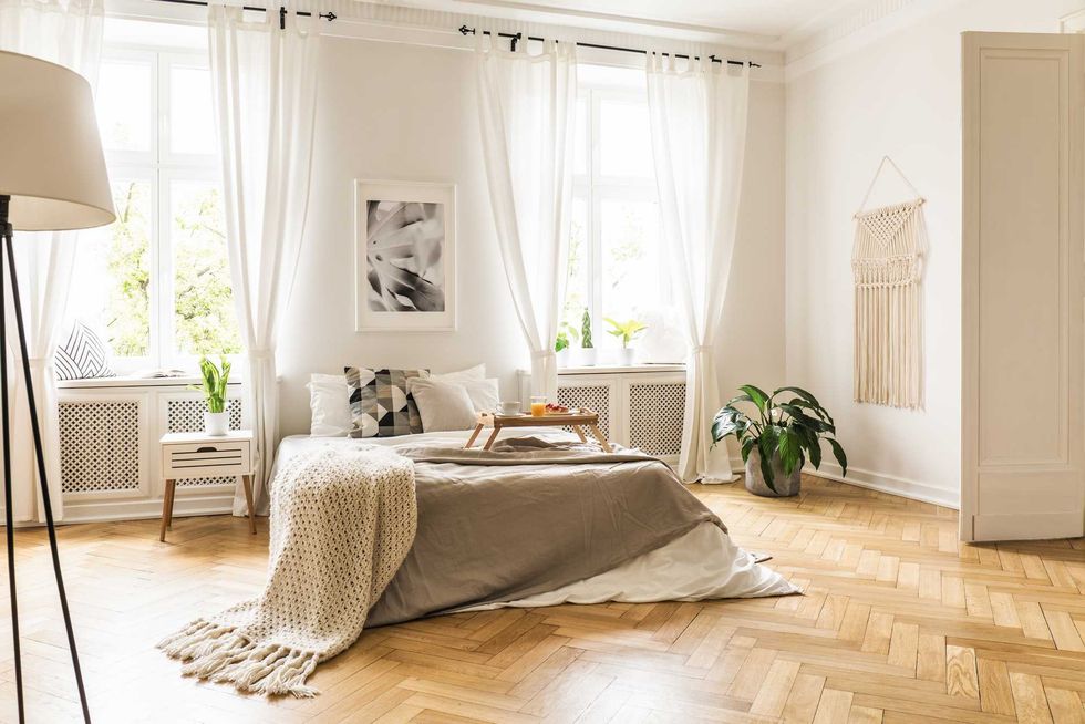 Framed poster on a white wall above a cozy double bed with beige sheets and breakfast tray in a spacious, sunny bedroom interior