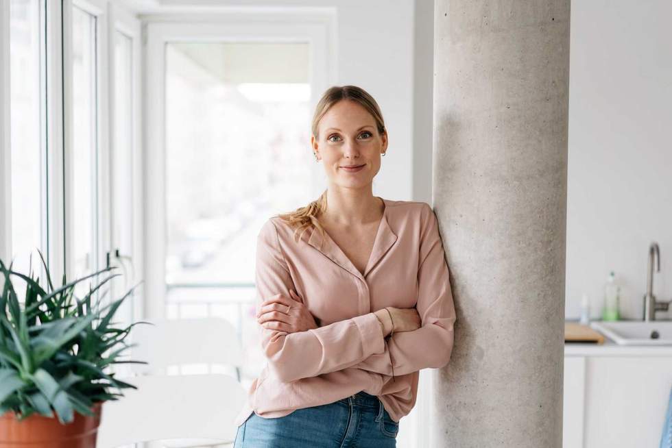 Friendly smiling young woman relaxing in an apartment kitchen leaning against an interior pillar with crossed arms looking at camera in a high key portrait with copyspace