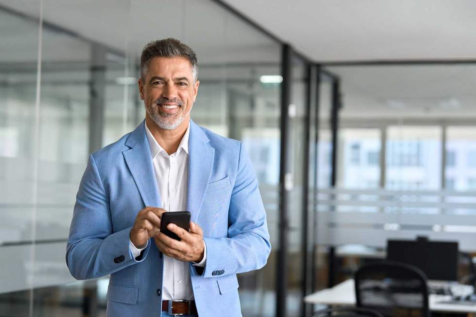 Happy mature business man investor wearing suit standing in office using mobile phone. 50 year old smiling businessman professional holding smartphone looking at camera at work. Portrait, copy space.
