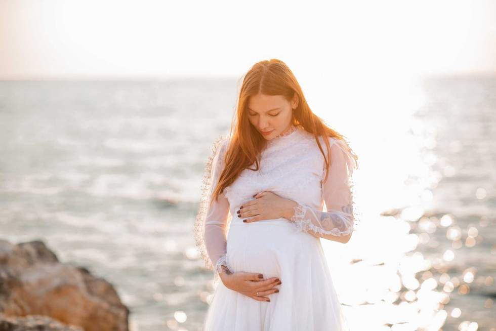 Happy pregnant woman 20-24 year old wearing white vintage dress standing over sea and sunset background close up. Maternity. Motherhood.