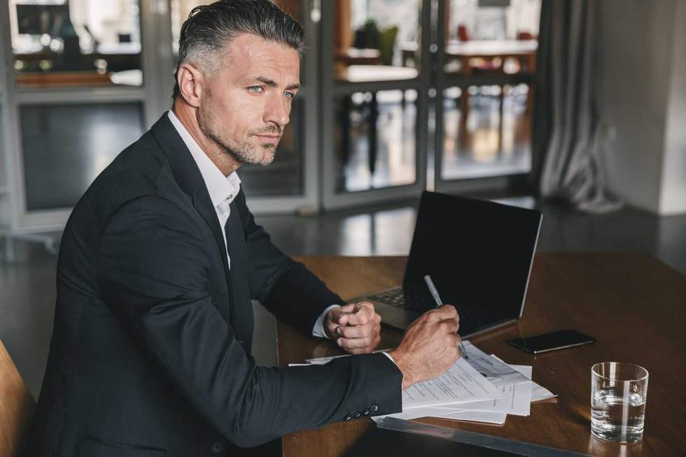 Image of concentrated confident businessman 30s wearing white shirt and black suit sitting at table in office during work with documents and laptop