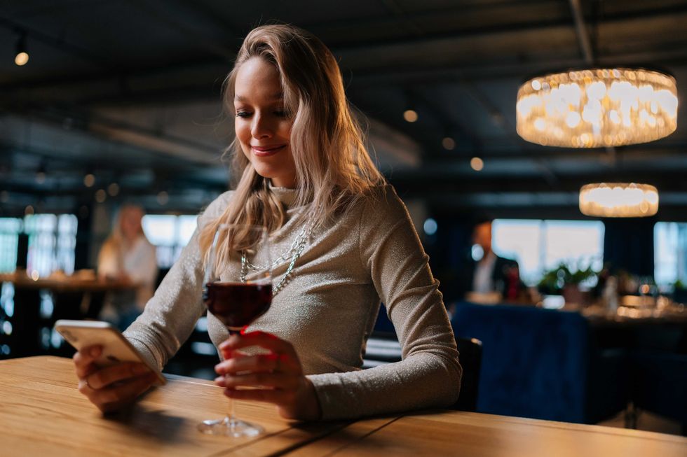 Low-angle view of smiling blonde young woman using smartphone, typing online message sitting at table holding in hand glass of red wine at restaurant. Happy pretty lady chatting with boyfriend.