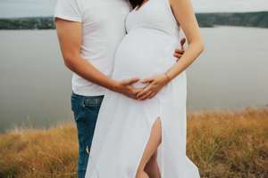 Man and his beautiful pregnant wife in white dress are hugging and standing on the hill near the lake.
