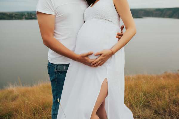 Man and his beautiful pregnant wife in white dress are hugging and standing on the hill near the lake.