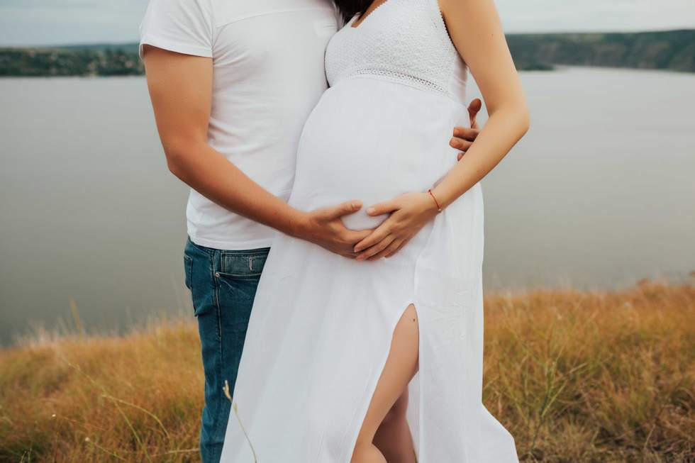 Man and his beautiful pregnant wife in white dress are hugging and standing on the hill near the lake.