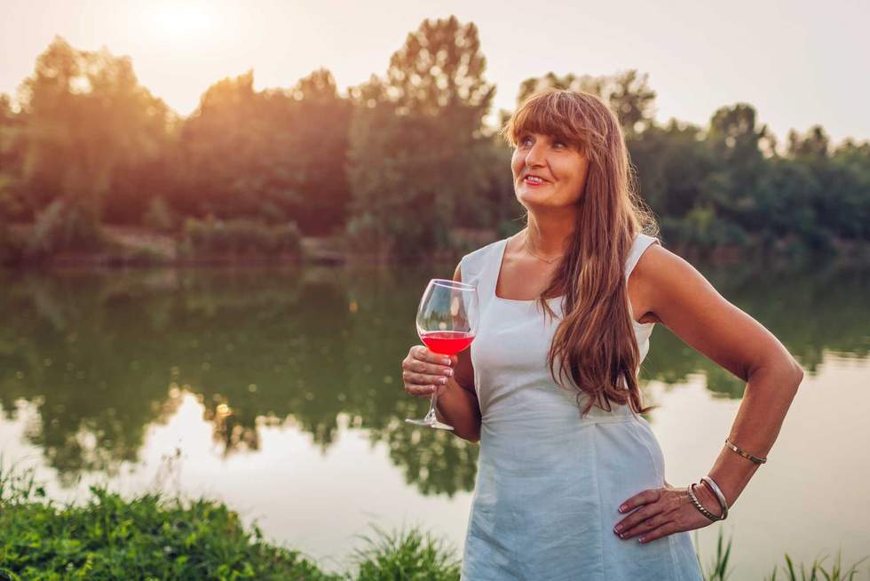 Mature woman enjoying glass of wine by river in autumn park. Senior woman admires landscape having drink
