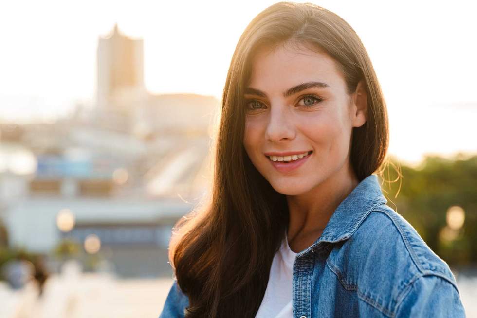 Portrait of a beautiful smiling young brunette woman standing outdoors with cityscape background, posing
