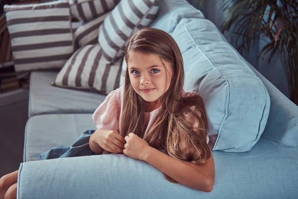Portrait of a smiling little girl with long brown hair and piercing glance, lying on a sofa at home.