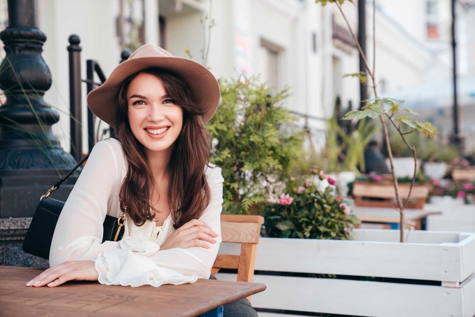 Portrait of beautiful young woman in hat. Model at coffee shop. Female outdoors in sunny day. In white blouse. Cheerful and happy. Waiting for an order at terrace cafe, looks great