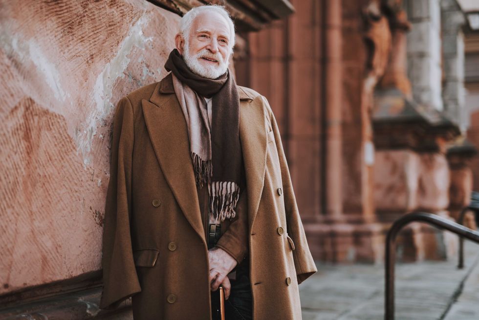 Portrait of old bearded man in coat and scarf holding walking stick while standing on the street. He is looking away and smiling