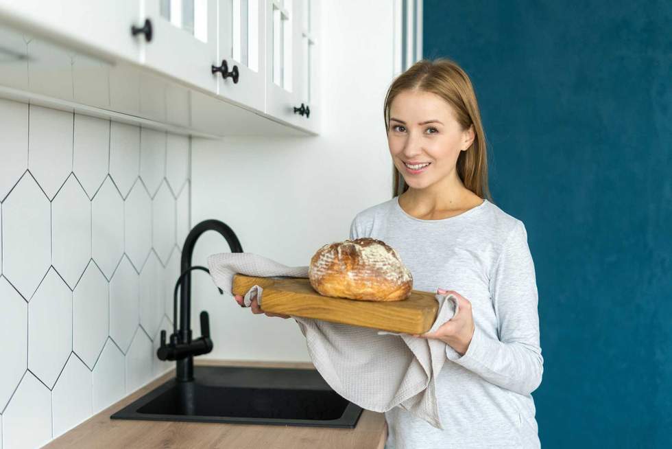 Portrait of smiling young lady holding freshly baked bread. Cooking traditional homemade food in kitchen. Bakery concept. Culinary hobby for women