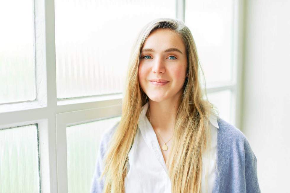 Portrait of young pretty 20 year old girl posing next to big window, looking straight at camera