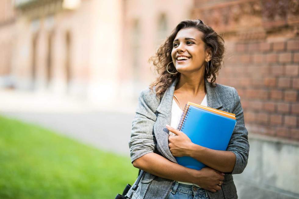 Pretty cheerful student smiling at camera carrying notebook on campus at college