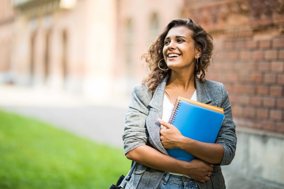 Pretty cheerful student smiling at camera carrying notebook on campus at college