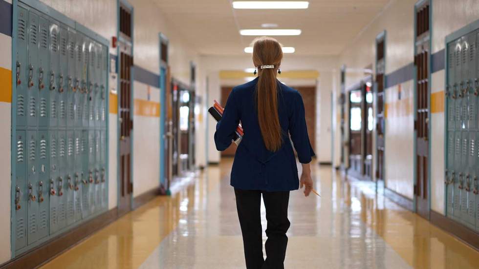 Rear view of teacher walking down a hallway in an empty school holding books showing emptiness.