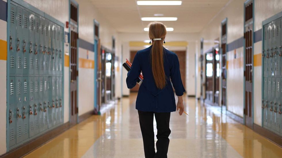 Rear view of teacher walking down a hallway in an empty school holding books showing emptiness.