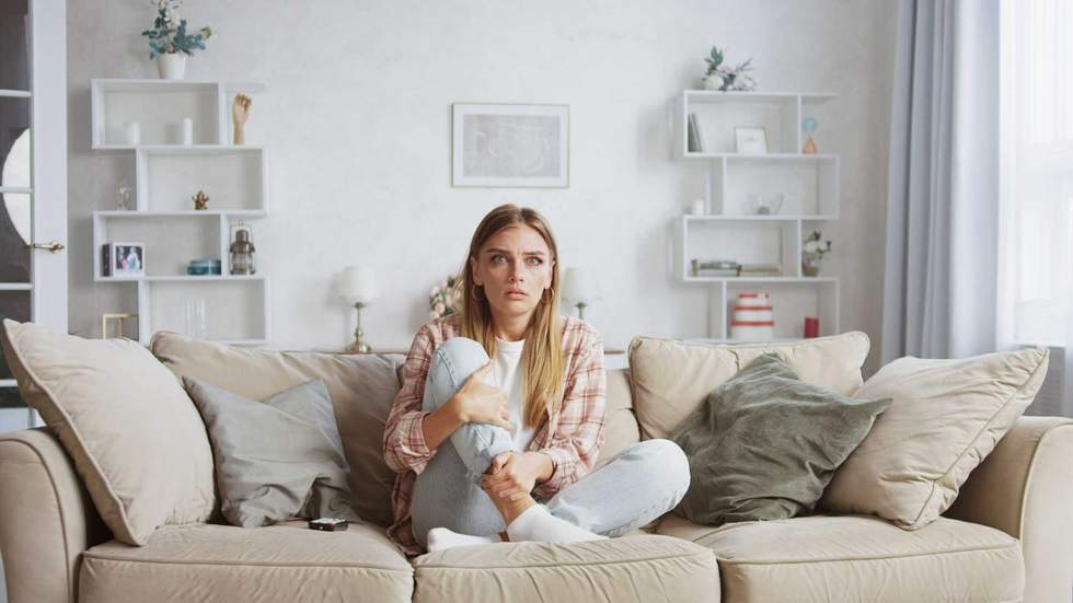 Sad woman is sitting on comfortable sofa with crossed legs and hugging her knees, she is crying while watching a drama movie or tv show in her living room