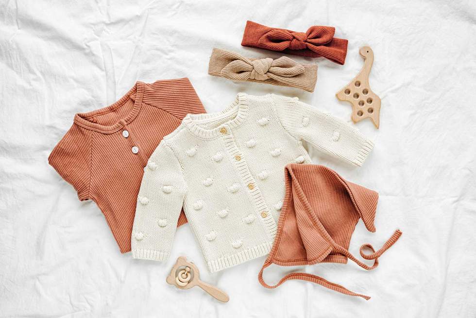 Set of baby rompers, hat, hairband and knitted jumper on white bed. Fashion baby clothes and accessories. Flat lay, top view