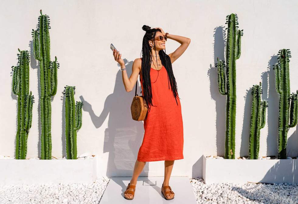 stylish smiling pretty woman with african braids on sunny summer vacation in colorful outfit wearing orange dress and sunglasses posing against white wall with cactuses in Mexico, using smartphone