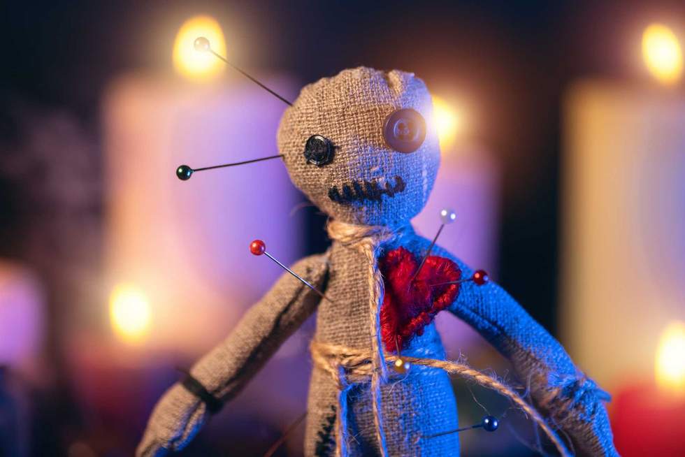 Voodoo doll studded with needles with pierced red rag heart against background of burning candles. Eerie or spooky magical esoteric ritual.