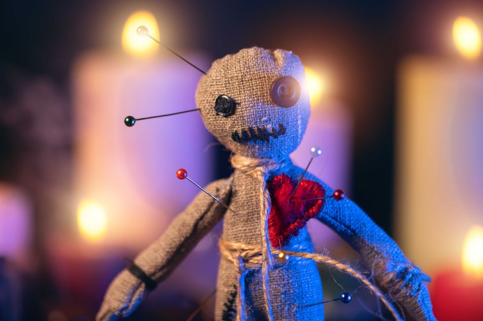 Voodoo doll studded with needles with pierced red rag heart against background of burning candles. Eerie or spooky magical esoteric ritual.