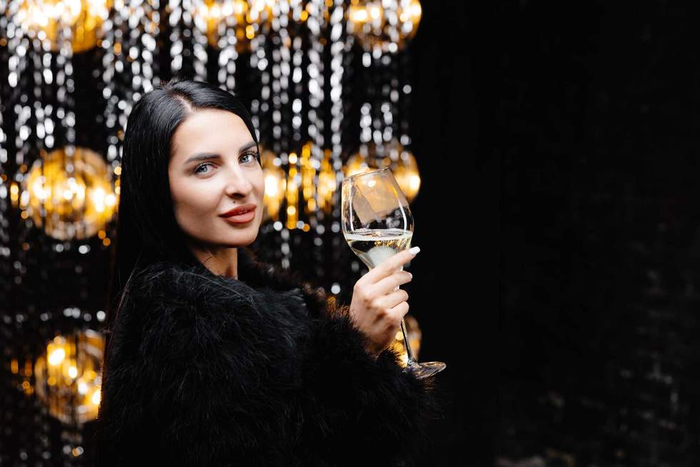 Young happy woman in trendy evening fur coat looking at camera and smiling with glass of champagne.