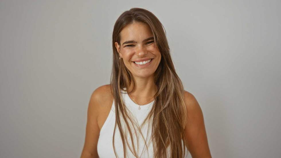Young hispanic woman smiling, standing against isolated white background, wearing white sleeveless top, brunette hair over her shoulders, exuding confidence and beauty in a relaxed setting