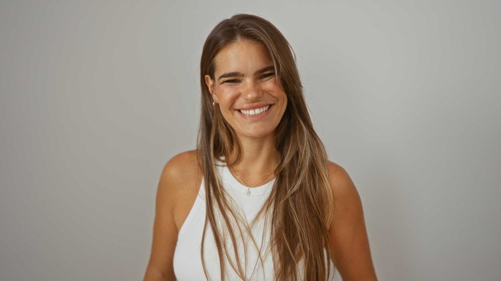 Young hispanic woman smiling, standing against isolated white background, wearing white sleeveless top, brunette hair over her shoulders, exuding confidence and beauty in a relaxed setting