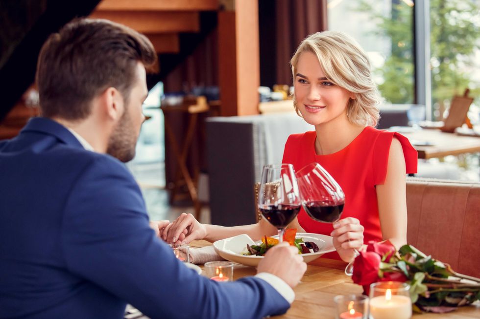 Young man and woman on date in restaurant sitting at table having lunch holding glasses drinking red wine cheers holding hands looking at each other passionate.
