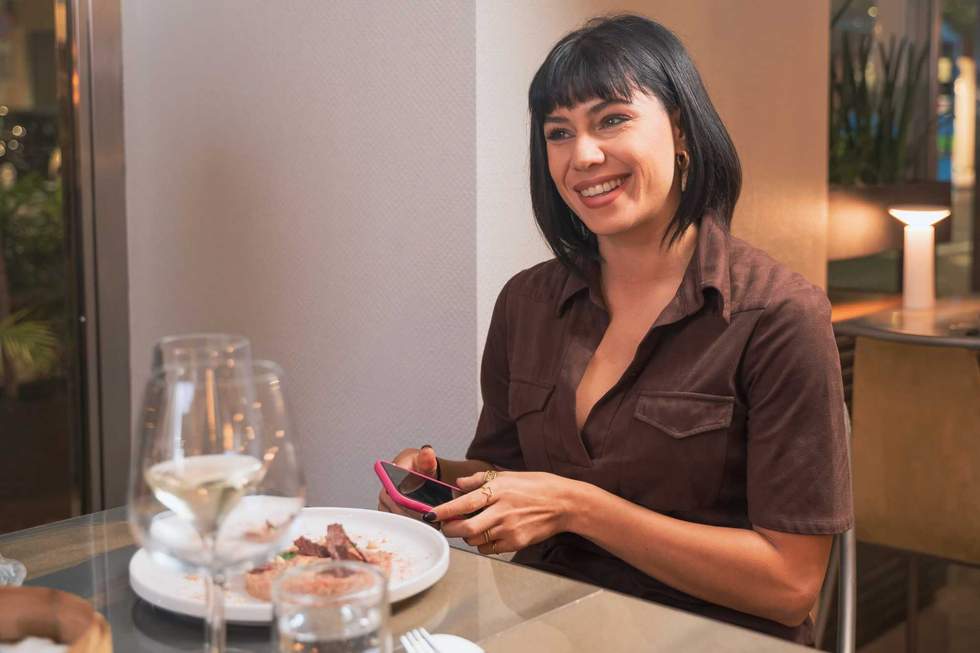Young professional woman using smartphone during dinner at upscale restaurant, smiling while checking social media