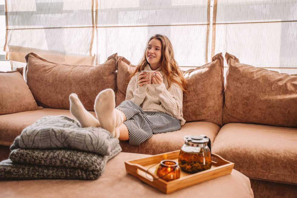 Young smiling woman sitting on sofa and looking up while drinking hot tea. Young millennial woman thinking at home in leisure time. Happy girl relaxing at home on bright winter morning