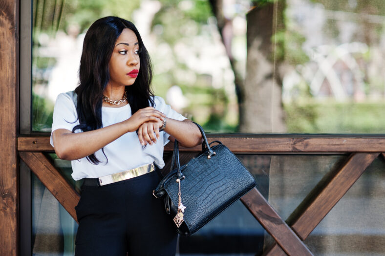 Stylish african american business woman with handbag on streets of city and looking at watches.