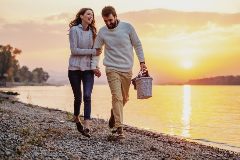 Feliz pareja de moda caucásica enamorada tomados de la mano y caminando por la costa cerca del río. Hombre sosteniendo la canasta de picnic. En el fondo está el atardecer.