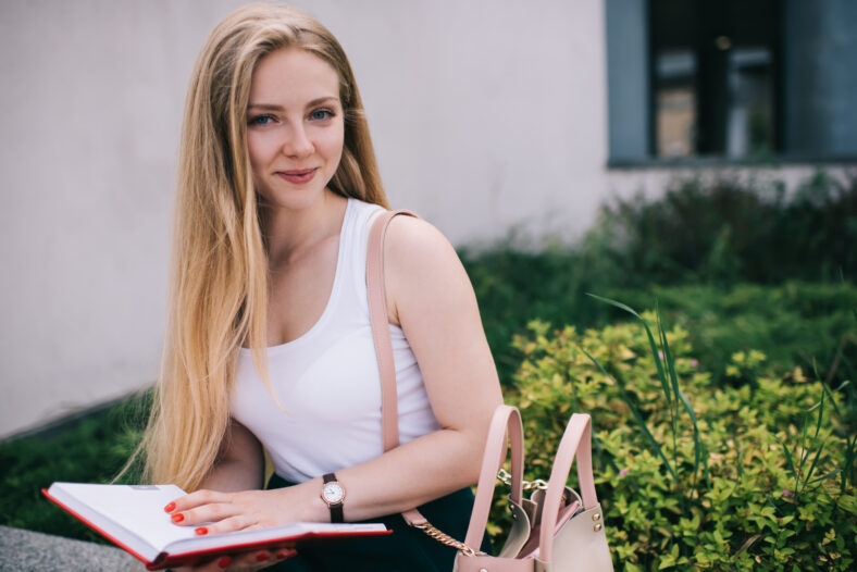 Content smiling blonde female with long hair and pink bag in sleeveless white shirt and green skirt reading book in red cover in street