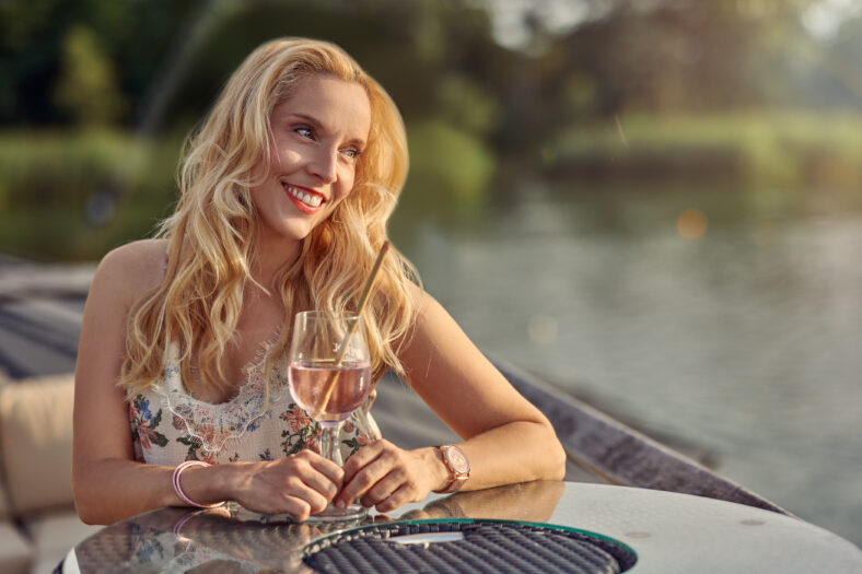 Happy young woman relaxing with a cocktail for sundowners seated at an open air table on a wooden deck overlooking calm water looking aside with a smile of pleasure enjoying a warm summer evening