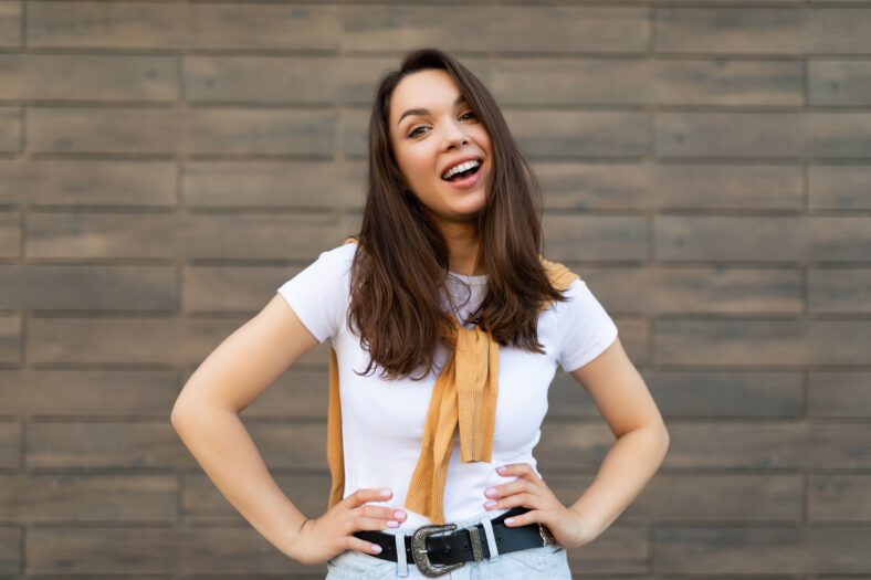 Portrait of young beautiful positive happy brunette woman standing against brown wall in the street and wearing stylish outfit.
