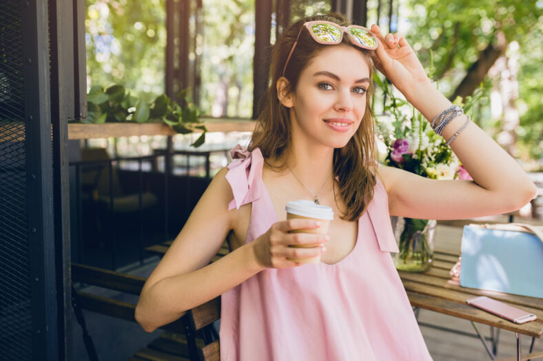 portrait of young smiling happy pretty woman with sitting in cafe drinking coffee, summer fashion outfit, hipster style, pink cotton dress, trendy apparel accessories