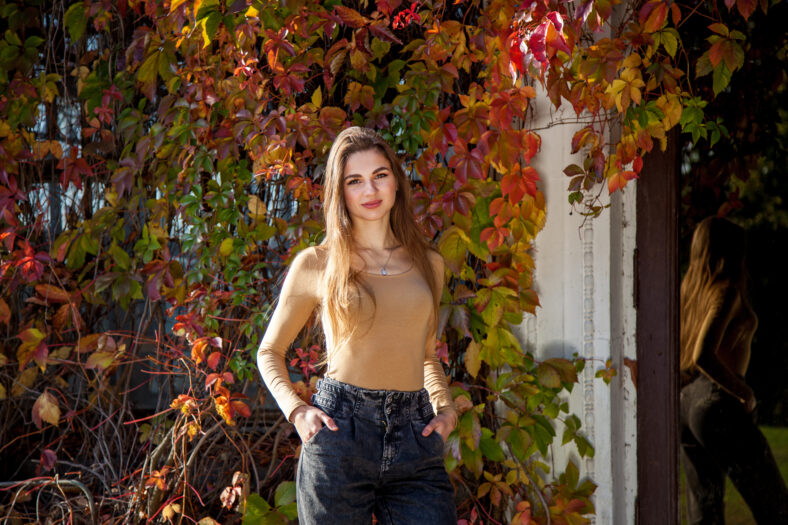 A girl with long hair in a bodysuit and jeans posing in an autumn park against the background of a wall overgrown with wild grapes. Girls in the park on the background of wild grapes