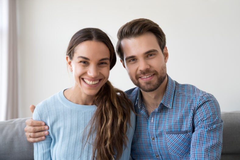Headshot portrait of happy smiling millennial couple sitting on sofa at home embracing looking at camera, young man and woman making video call communicating online or recording vlog together
