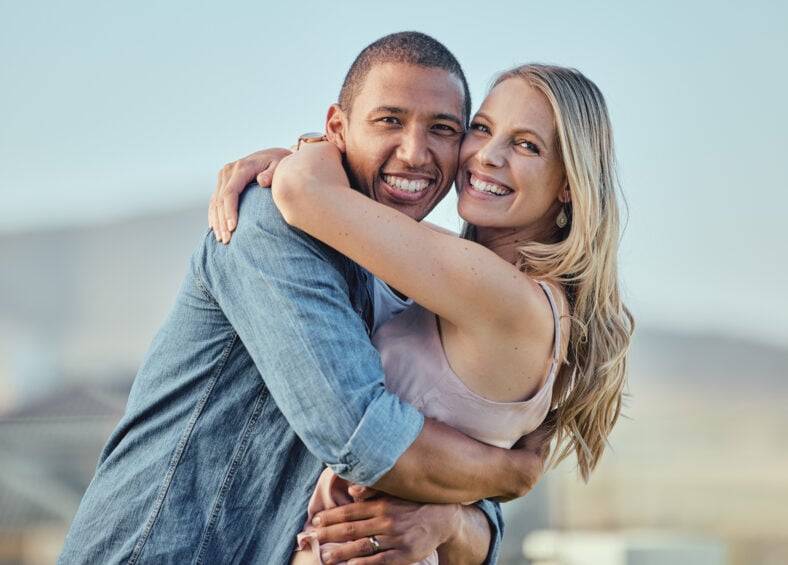 Interracial, happy couple and smile portrait hug of people in summer with happiness or love. Couple, outdoor and smiling for marriage, care or vacation gratitude in sunshine together feeling calm.