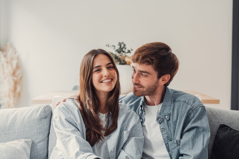Portrait of young 20s just married couple in love posing photo shooting seated on couch in modern studio apartments, concept of capture happy moment, harmonic relationships, care and sincere feelings