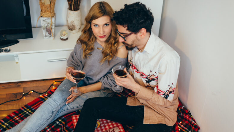 Young couple toasting with cocktail glasses at home in living room, girl looking at camera